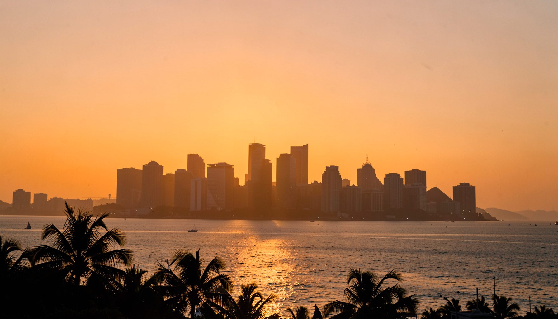 海口城市天际线在夕阳下的剪影，金色余晖映照海面，椰树轮廓点缀前景